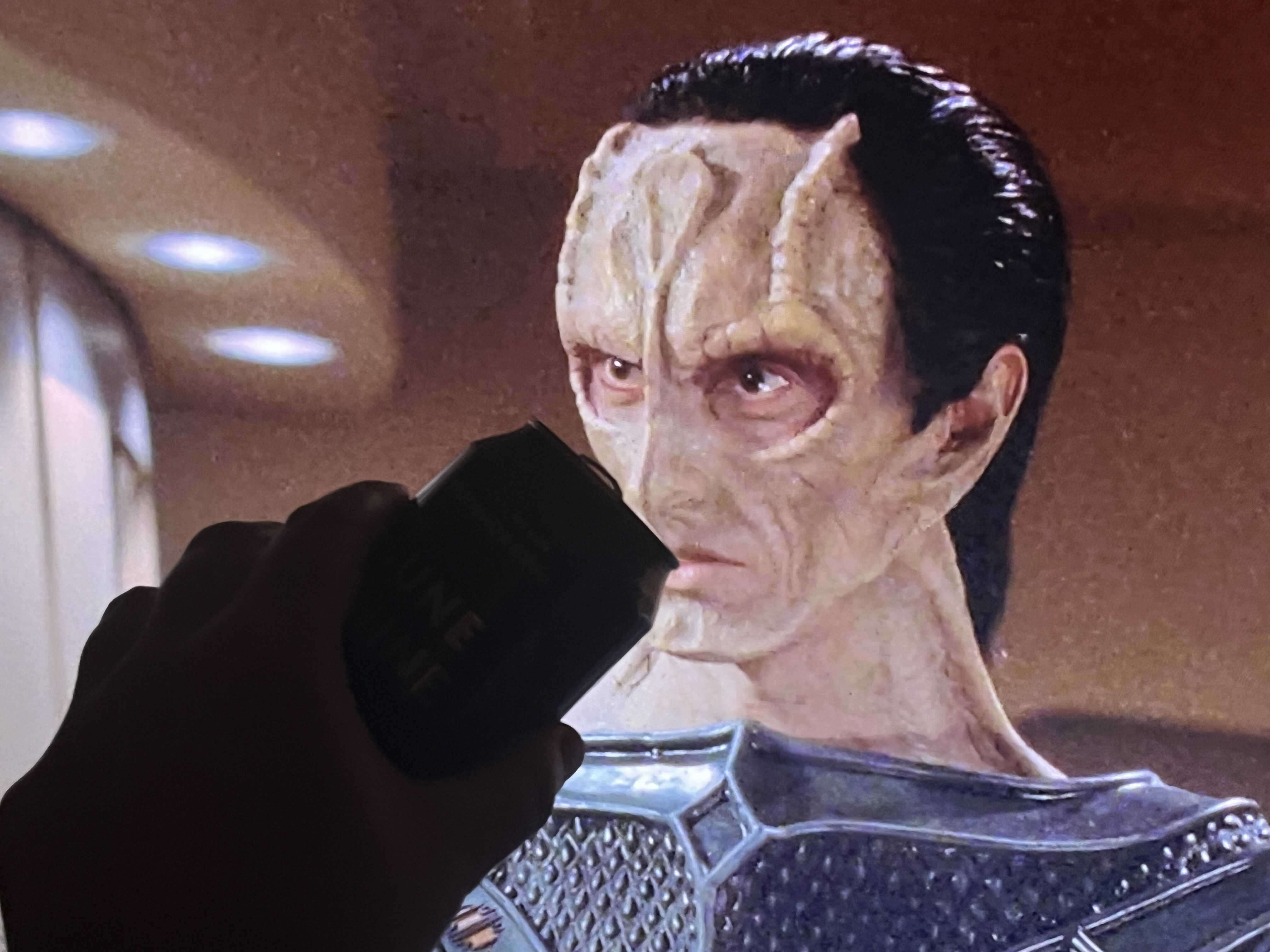 A Star Trek Cardassian with slicked-back hair and dark shoulder garment stands in an interior space with beige walls. A hand holding an beer near the figure’s mouth on the tv screen is visible.