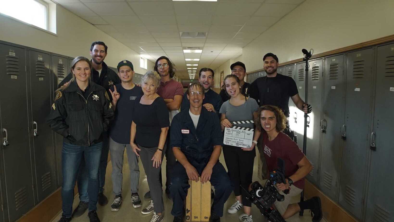 The image depicts the crew of the short film Custodial Arts, standing as a group in hallway with lockers on either side. They are holding the items connected to their jobs, like a boom microphone, camera, slate, and costumes for the actiors. They are smiling and happy to have wrapped the film!